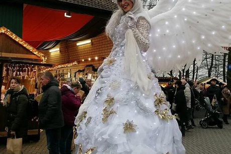 Die als Engel verkleidete Stelzenläuferin hat sich für den Fotografen in Positur geworfen und lächelt in die Kamera. Im Hintergrund erkennt man beleuchtete Weihnachtsmarktbuden.