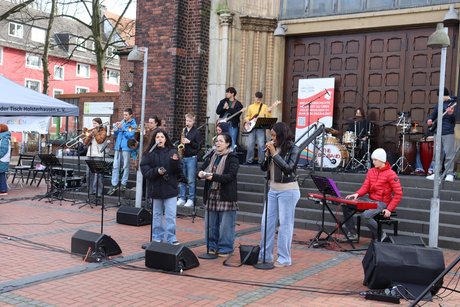 Jugendliche stehen auf den Stufen einer Kirche mit Instrumenten oder vor Mikrophonen und singen.