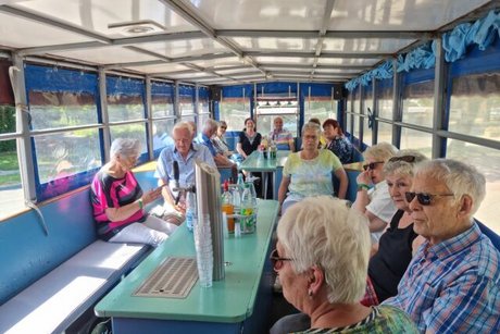 Eine Gruppe Menschen sitzt auf Bänken um Tische herum auf einem langen Planwagen.