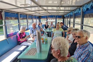 Eine Gruppe Menschen sitzt auf Bänken um Tische herum auf einem langen Planwagen.