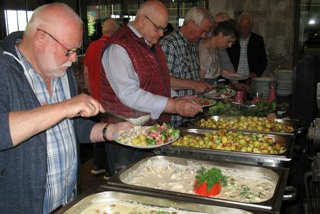 Menschen stehen an einem Buffet und füllen sich Teller mit verschiedenen Gerichten, die dort in Edelstahlbehältern angerichtet sind.
