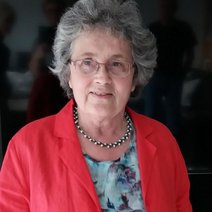 Abgebildet ist die Schriftführerin Ulrike Nierhaus. Sie trägt einen roten Blazer über einer geblümten Bluse, hat kurze, leicht gelockte graue Haare und ist Brillenträgerin