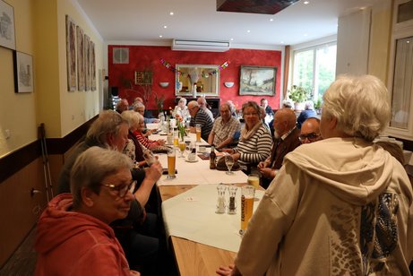 Menschen sitzen an einem langen Tisch und h&ouml;ren einer Frau zu, die steht.