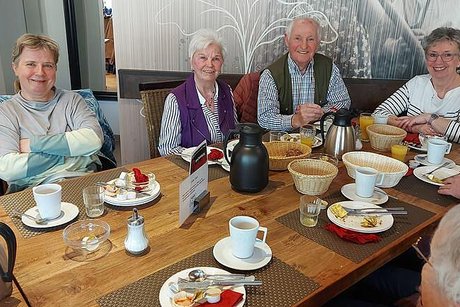 Drei Frauen und ein Mann sitzen gemeinsam ums Eck an einem gedeckten Frühstückstisch und lächeln in die Kamera.