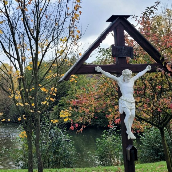 G&ouml;ckenteich mit Blick auf die Nikomedeskirche. Vorne im Bild ein dunkles Holzkreuz mit heller Jesusfigur. Links im Bild, 2 schwarze Schw&auml;ne am grasen.