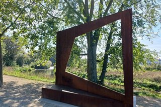 Ein rostiger, mannsgroßer Rahmen steht am Rand eines von Bäumen gesäumten Weges. Dahinter ist eine grasbewachsene Brachlandschaft.