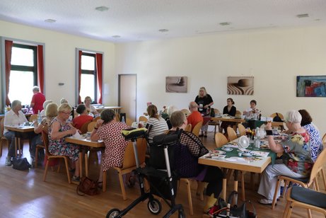 Eine Frau steht und spricht zu den Teilnehmern an drei langen Tischen.