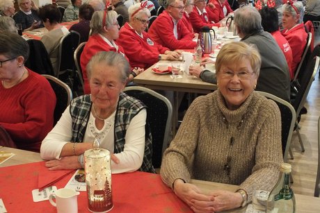 Zwei Frauen sitzen an einem Tisch. Dahinter sitzen viele andere Menschen an langen Tischen.