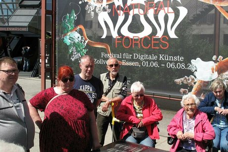 Die Reisegruppe steht um einen barrierefreien Geländeplan aus Bronzeguss herum. Im Hintergrund wirbt ein Plakat für die Ausstellung "New Natural Forces Now" zum Thema Digitale Künste.