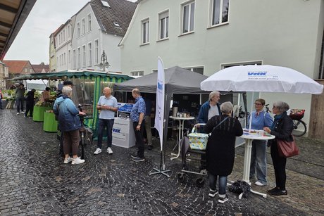 Marktstand Der Stand ist aufgebaut. Vorne rechts unterm Sonnenschirm ist ein Stehtisch. Hier stehen 4 Personen am Tisch und unterhalten sich. Dahinter ist ein Pavillion aufgebaut.