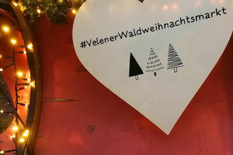 Ein großes, weißes Herz hängt an der Rückwand einer mit Tannengrün und Lichterkette geschmückten, roten Kutsche. Die Aufschrift auf dem Herz lautet #velenerweihnachtsmarkt