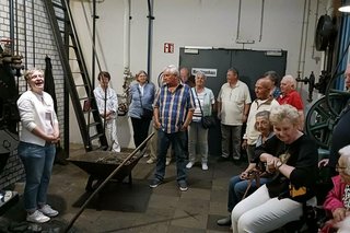 Eine Gruppe Menschen steht oder sitzt auf Rollatoren in der Eingangshalle des Textilmuseums Bocholt und hört zu, was eine Frau links im Bild erzählt.