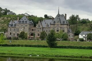 Panoramafoto der Mosel mit einem herrschaftlichen Anwesen und Nebengebäuden und im Hintergrund sich erhebenden Weinbergen