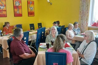 Menschen sitzen gemeinsam an Tischen im Restaurant der Straußenfarm. An der Seite des Raumes hängen Bilder von Straußen.