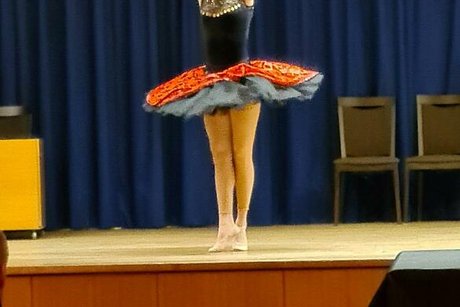 Eine junge Frau in schwarzem Kostüm mit schwarz-rotem Tutu steht in Tanzpose mit Taburin in der Hand auf der Bühne.
