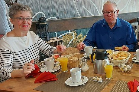 Ein Mann und eine Frau sitzen gemeinsam ums Eck an einem reichhaltig gedeckten Frühstückstisch und schauen in die Kamera.