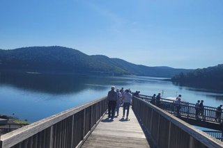 Die Sonne strahlt vom wolkenlosen Himmel auf einen langen Holzsteg, der Richtung See führt. Am Ende des Stegs sind entfernt einige Menschen zu erkennen.