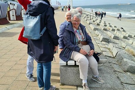 Einige Menschen stehen oder sitzen auf einer Bank an der Uferpromenade von Norderney. Im Hintergrund sieht man Wellen, die ans Ufer schlagen.
