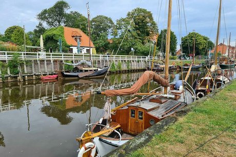 Alte Fischkutter liegen rechts und links am Ufer einer Gracht vertäut. Im Hintergrund sieht man das rote Dach eines Hauses zwischen Bäumen.