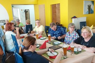 Menschen sitzen gemeinsam an Tischen im Restaurant der Straußenfarm. An der Wand gemalt sind Straußen.