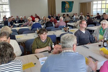 Viele Menschen sitzen gemeinsam in einem Saal an langen Tischen.