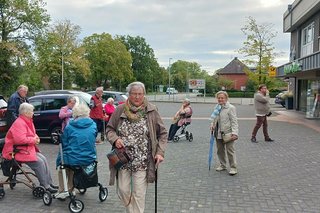 Eine Gruppe Menschen wartet auf einem Parkplatz. Einige davon sitzen auf Rollatoren.