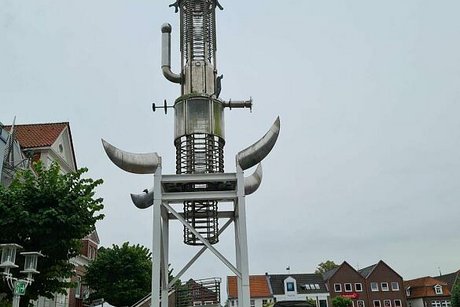 Auf dem Marktplatz von Aurich ragt der Sous Turm in den wolkenverhangenen Himmel. Die etwa 25 Meter hohe Skulptur aus stahlverkleidetem Plexiglasspiralen mit Hörnern und Schornstein erinnert entfernt an einen Tauchsieder.