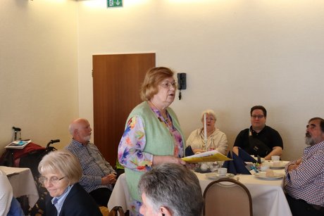 Eine Frau liest etwas von einem Zettel ab, um die Mitglieder zu begr&uuml;&szlig;en.