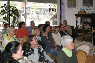 Viele Menschen sitzen gemeinsam in einem Raum an Tischen und hören einem Vortrag zu.