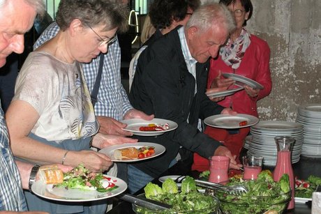 Menschen stehen an einem Salat-Buffet und füllen sich Teller mit verschiedenen Salaten, die dort in Glasschüssel angerichtet sind.