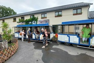 Menschen steigen in eine blau-weiß bemalte Bimmelbahn, die im Innenhof der Straußenfarm Halt macht.