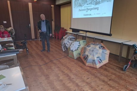 Schirmmacher Willy Schüffler hält einen Vortrag über seinen Beruf und die Geschichte des Schirms. Herr Schüffler, von Beruf Schirmmacher, hält einen Vortrag und steht vor einer Leinwand.