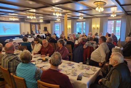 Info Nachmittag Mitglieder sitzen an den Tischen und hören gespannt den Vortrag der Referentin. Referentin sitzt vorne am Tisch und zeigt eine Power Präsentation.