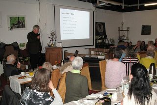 Viele Menschen sitzen gemeinsam in einem Raum an Tischen und hören einem Mann zu, der mit Mikrofon vor einer Powerpoint-Präsentation steht.