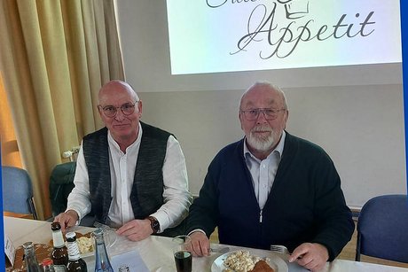 Zwei Männer sitzen vor Tellern mit Schnitzeln und Kartoffelsalat an einem Tisch. Hinter ihnen leuchtet ein Beamer-Bild an der Wand mit dem Spruch: Guten Appetit.