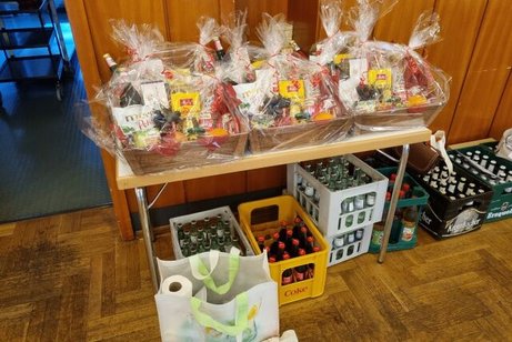 Verschiedene in Zellophan verpackte Präsentkörbe stehen auf einem Tisch. Darunter und daneben stehen Kästen mit verschiedenen Getränken.