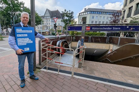 Ein Mann steht mit einem Schild, vor einer gesperrten Rolltreppe. Auf dem Schild wir nach mehr Barrierefreiheit gefordert.