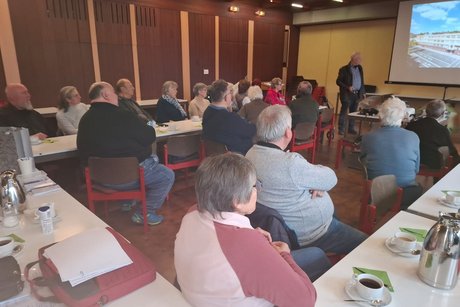 Schirmmacher Willy Schüffler hält einen Vortrag über seinen Berufbild und die Geschichte des Schirms. Mehrere Mitglieder sitzen an langen Tischen und verfolgen den Vortrag.