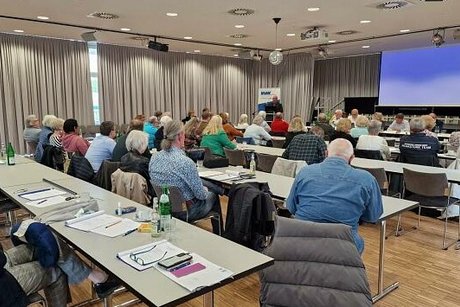 Menschen sitzen an Tischen und folgen einem PowerPoint Vortrag
