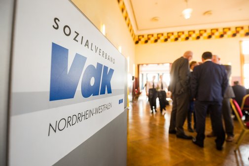 Blick in einen Saal voller Menschen an Stehtischen. Links im Bild ein VdK-Logo an einer grauen Stellwand.