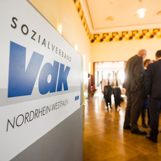 Blick in den Saal Blick in einen Saal voller Menschen an Stehtischen. Links im Bild ein VdK-Logo an einer grauen Stellwand.