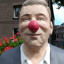 Die Skulptur Alltagsmensch in Straelen ist ein Mann mit blondem Haar, der im Gesicht eine rote Clowns-Nase hat, angezogen mit grauer Jacke und weißes Hemd.