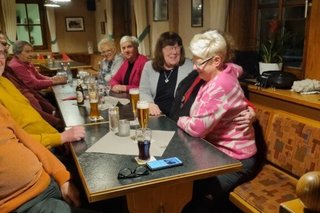 Eine Gruppe Menschen sitzt gemütlich in einer Gaststube an einem langen Tisch und redet und lacht.