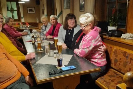 Eine Gruppe Menschen sitzt gemütlich in einer Gaststube an einem langen Tisch und redet und lacht.