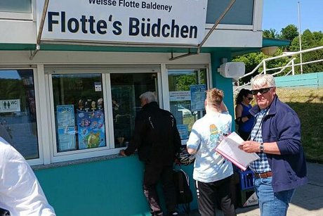 Ein Mann mit Schiebermütze steht in einer Schlange an einem Kiosk, dass mit "Flottes Büdchen" beschriftet ist.