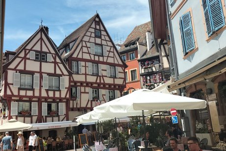 Im Vordergrund an der rechten Bildseite ist ein Straßencafé mit Sonnenschirmen zu sehen; im Hintergrund zeigen sich schöne Fachwerkfassaden.