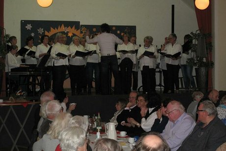 Ein Chor mit Chorleiter steht auf einer Bühne und singt. Vor der Bühne sitzen Menschen an einem weihnachtlich geschmücktem Tisch.