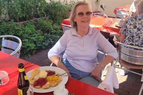 Eine Frau sitzt an einem Tisch mit einer großen Waffel und einem Kännchen Kaffee vor sich und lächelt in die Kamera.