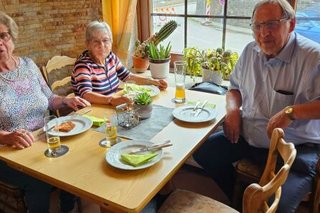 Zwei Frauen und ein Mann sitzen gemeinsam an einem gedeckten Holztisch vor einem Fenster.