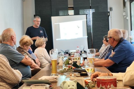 Mitglieder sitzen an einem langen Tisch und h&ouml;ren dem stehenden Referenten zu, der neben einer Leinwand steht.